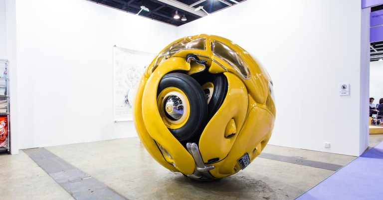 Yellow 1953 Volkswagen Beetle compressed into a giant sphere, displayed in a white gallery booth