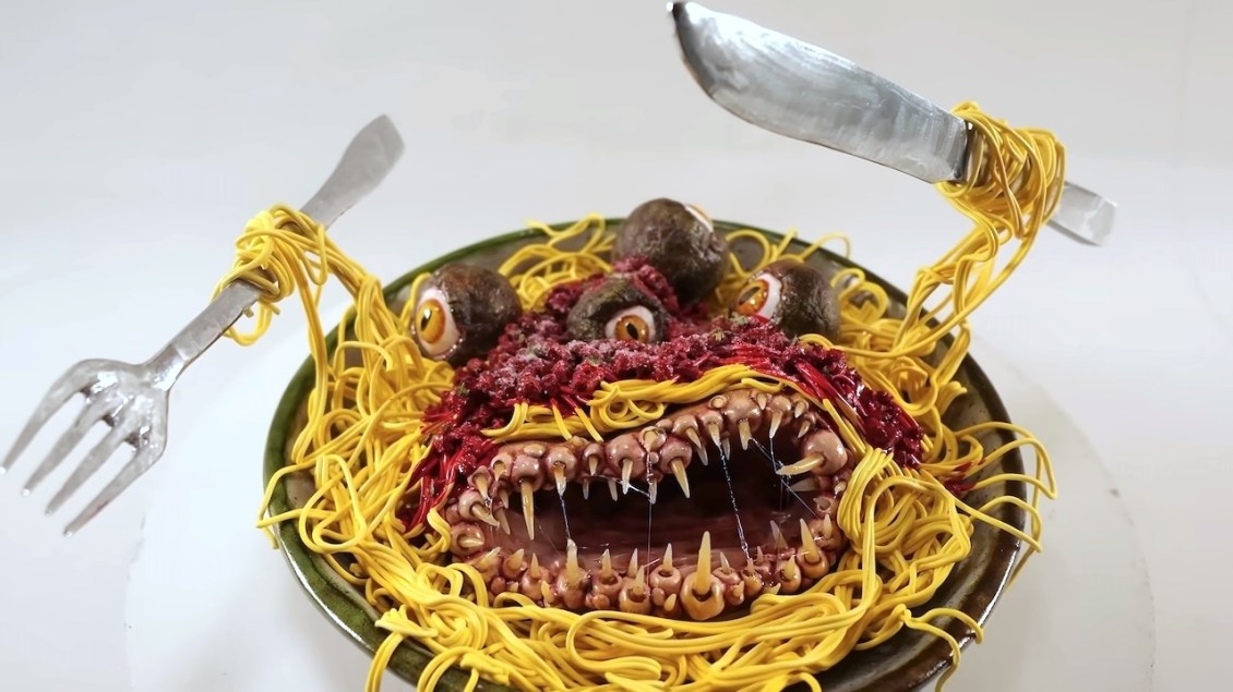 Clay sculpture of an evil spaghetti monster in a bowl with sharp teeth, eyeballs, and noodle arms holding a fork and knife